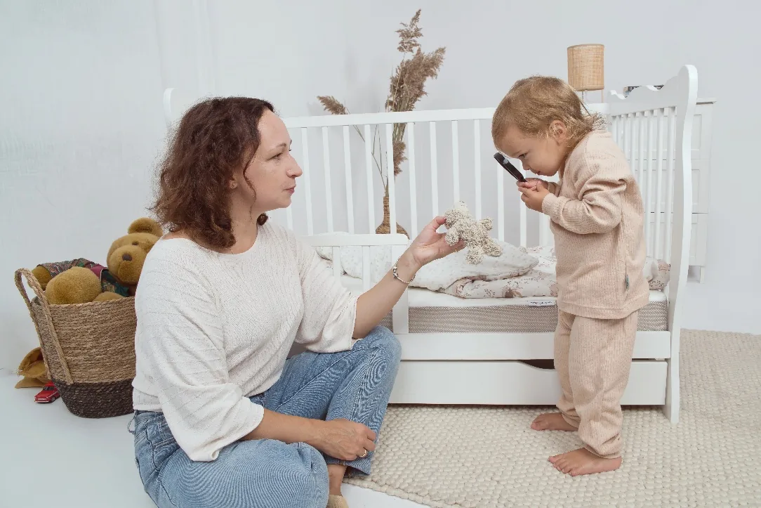 Mama przy łóżeczku obserwuje dziecko, które przez lupę ogląda materac